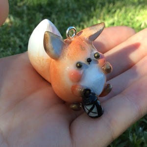 Puede incluir: Figurita de zorro hecha a mano con colores naranja, blanco y marr&oacute;n, sosteniendo una linterna negra. El zorro tiene un lazo plateado para colgar. Es un objeto decorativo caprichoso.
