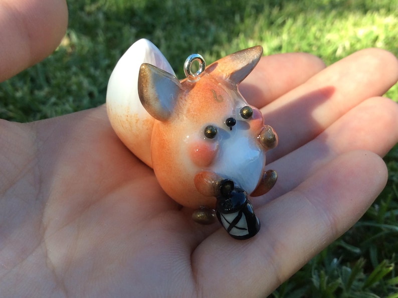 Puede incluir: Figurita de zorro hecha a mano con colores naranja, blanco y marr&oacute;n, sosteniendo una linterna negra. El zorro tiene un lazo plateado para colgar. Es un objeto decorativo caprichoso.