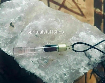 Op de afbeelding: Een heldere glazen flacon met een gouden dop en een donkerrode substantie erin, opgehangen aan een zwart koord. De flacon rust op een groot, ruw, lichtgekleurd kristal. De tekst "SpiritualstaffShop" is zichtbaar.