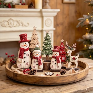 Puede incluir: Una colección de figuras navideñas de madera sobre una bandeja redonda, que incluye dos muñecos de nieve con sombreros y bufandas rojas, dos árboles de Navidad estilizados en verde y rojo, un árbol de madera natural y un reno con lazo verde.