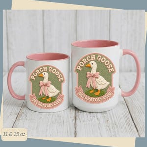 May include: Two white ceramic mugs with pink interiors and handles, featuring a graphic of a white goose wearing a pink bow on a green background. The text reads Porch Goose Decorators Club. The mugs are available in 11 and 15 ounce sizes.