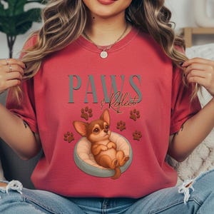 May include: Red t-shirt with the text "PAWS & Reflect" and a cartoon chihuahua in a bed. The design includes paw prints. The shirt is a solid color.