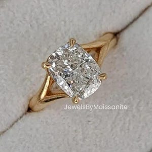 May include: A gold ring with a large, square-cut, clear gemstone. The ring is set in a four-prong setting with a split shank design. The ring is displayed in a gray velvet ring box. Text on the box reads "JewelsByMoissanite".