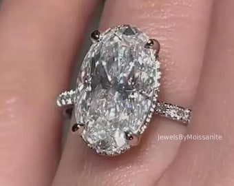 May include: A diamond ring featuring an oval-cut central stone, surrounded by a halo of smaller diamonds. The band also has small diamonds. The ring is set in silver. The text "JewelsByMoissanite" is visible.