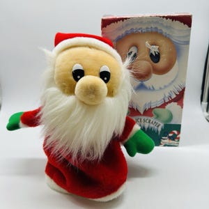Könnte beinhalten: Eine Plüschpuppe des Weihnachtsmanns mit rotem Anzug, Mütze und grünen Fäustlingen. Die Puppe hat einen weißen Bart und eine runde, beige Nase. Im Hintergrund befindet sich eine Schachtel mit einer Weihnachtsmann-Illustration. Auf der Schachtel steht "ICE SCRAPER".