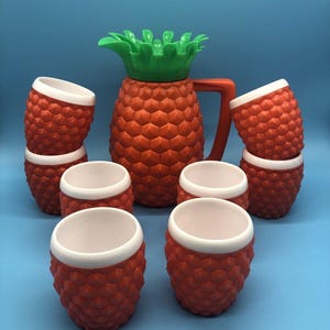 May include: A set of orange pineapple-shaped drinkware, including a pitcher and several matching cups. The pitcher has a green top and a red handle. The cups are orange with white rims. The set is arranged on a blue background.