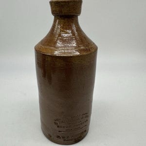 May include: Brown stoneware bottle with a cylindrical body and a short, flared neck. The bottle has a textured surface and appears to be antique. The base of the bottle has embossed text. The bottle is likely a vintage piece.