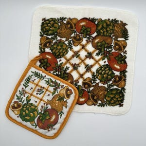 May include: A white kitchen towel and pot holder set with a vegetable pattern. The design includes artichokes, tomatoes, and mushrooms in shades of green, brown, and orange. The pot holder has an orange border.