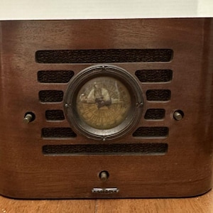 May include: A dark brown wooden radio with a rounded rectangular shape. It features a central circular dial with a metallic frame, horizontal speaker slats, and three small control knobs positioned on the front panel. The brand name Philips is visible.