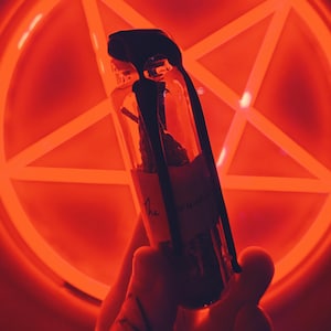 Może przedstawiać: Przezroczysta szklana butelka z czarną zakrętką i etykietą, trzymana na tle świecącego na czerwono pentagramu. Butelka zawiera suszone zioła. Tło jest jaskrawoczerwone, tworząc dramatyczną i mistyczną estetykę.