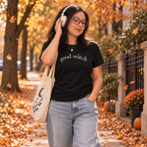Puede incluir: Una camiseta negra de manga corta con el texto blanco "good witch" impreso en el pecho. La prenda se combina con unos vaqueros de mezclilla azul claro y un bolso de tela color crema decorado con un gráfico negro de luna creciente y hojas.