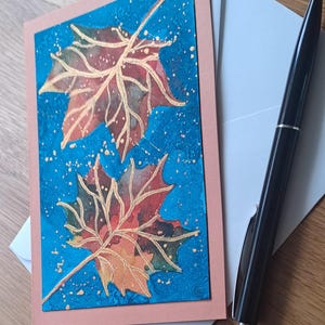Puede incluir: Tarjeta de felicitación con dos hojas de otoño con contornos dorados y detalles en acuarela en tonos rojos, naranjas y verdes sobre un fondo azul. La tarjeta tiene un borde melocotón, un sobre blanco y un bolígrafo negro.