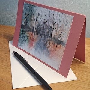 Può includere: Biglietto di auguri fatto a mano, color rosa antico, con un dipinto ad acquerello di un paesaggio. Una penna nera è appoggiata sulla busta bianca. Il dipinto raffigura alberi e il loro riflesso nell'acqua.