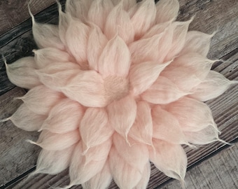 May include: A large, soft, light pink felt flower with multiple layers of petals. The flower is centered and has a textured appearance, with each petal tapering to a point. The background is a wooden surface.