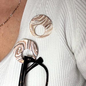 May include: A cream-coloured ribbed cardigan with a gold chain necklace and two circular brooches. The brooches feature a marbled design in beige and brown. Black eyeglasses are attached to one brooch.