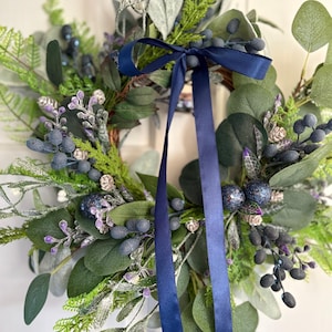May include: A circular wreath featuring green eucalyptus leaves, fern fronds, and frosted foliage. The arrangement is accented with clusters of dark blue berries, small glittery purple sprigs, and a long, smooth navy blue satin ribbon tied at the top.