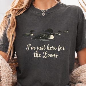Könnte beinhalten: Ein anthrazitfarbenes Kurzarm-T-Shirt mit einer Grafik von drei Eistauchern, die auf dem Wasser schwimmen. Unter den Vögeln steht der Text "I'm just here for the Loons" in einer cremefarbenen Schreibschrift. Das T-Shirt hat einen Rundhalsausschnitt.