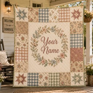 May include: A rectangular patchwork quilt featuring a central cream-coloured panel with a floral wreath and the text Your Name in brown script. The border consists of alternating squares with floral patterns, blue and white gingham, and star motifs.