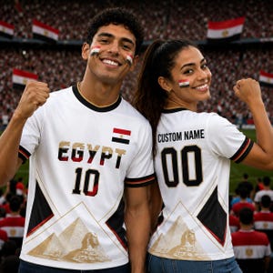 Könnte beinhalten: Ein weißes Sporttrikot mit dem Wort EGYPT in fetter Schrift, der Nummer 10 und einer kleinen ägyptischen Flagge. Das Design enthält schwarze und goldene geometrische Akzente mit einer Grafik der Pyramiden und der Sphinx am unteren Saum.