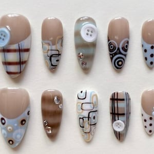 Könnte beinhalten: Eine Sammlung von zehn mandelförmigen Kunstnägeln mit verschiedenen Designs. Die Nägel haben eine neutrale Grundfarbe mit Akzenten wie Karomuster, Tupfen, geometrischen Mustern und Knöpfen. Einige Nägel haben kleine Strasssteine.