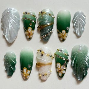 May include: A set of ten artificial nails in various designs. The nails feature a mix of green, white, and silver colors with floral and leaf accents. Some nails are adorned with rhinestones and gold-colored bands, creating an elegant and artistic look.