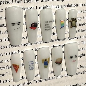 May include: A collection of ten white false nails, each decorated with distinct designs. The designs feature cartoon faces, a Roblox logo, a rainbow flag, a teddy bear, a pizza slice, and the text "eat, sleep, play roblox, repeat".