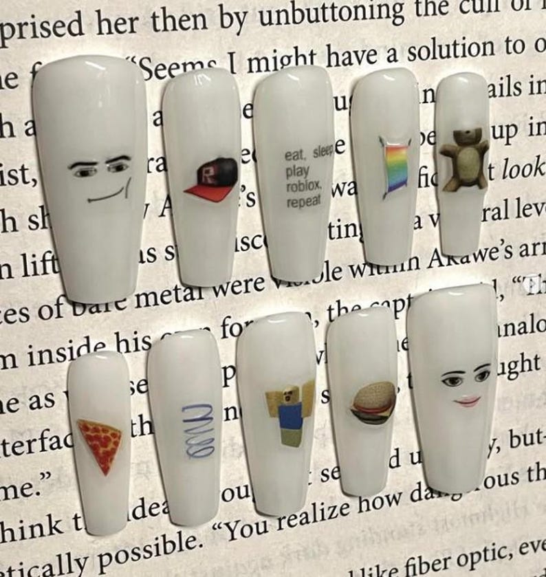 May include: A set of ten white artificial nails, each adorned with unique designs. The designs include cartoon faces, a Roblox logo, a rainbow flag, a teddy bear, a pizza slice, and the text "eat, sleep, play roblox, repeat".