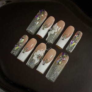 May include: A set of ten long, artificial nails with various designs. Some nails feature a nude base with white tips, while others have glitter and jewel embellishments. One nail has a white flower accent.