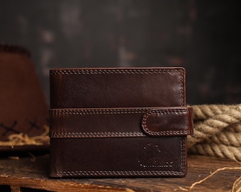 May include: A dark brown leather wallet with a stitched design and a snap closure. The wallet has a horizontal strap across the front. The wallet is sitting on a wooden surface.