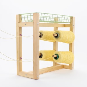 May include: A wooden yarn storage rack with a light green wire basket on top. The rack holds four large spools of yellow yarn. The rack is made of light-colored wood and has a simple, functional design.