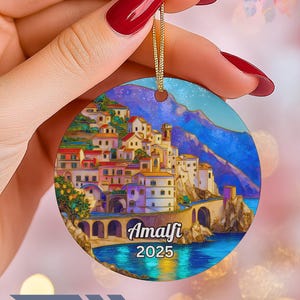 Pode incluir: Enfeite de Natal redondo com uma ilustração colorida de Amalfi, Itália, com edifícios, uma ponte e o texto "Amalfi 2025". O enfeite tem um cordão dourado para pendurar.