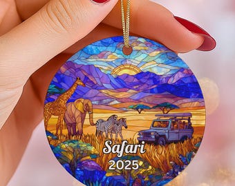 May include: Round ornament with a safari scene featuring a giraffe, elephant, zebras, and a safari vehicle. The ornament has a stained glass style with a sunset and the text "Safari 2025".