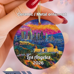 May include: A round ceramic or metal ornament featuring a vibrant, painterly depiction of the Los Angeles skyline at dusk, including the Griffith Observatory and the Hollywood sign, with the text Los Angeles 2026 printed in white at the bottom.
