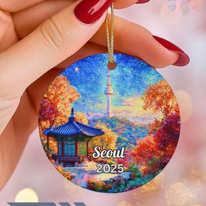 May include: Round ceramic ornament featuring a colorful illustration of Seoul, South Korea, with a pagoda and tower. The ornament has a gold string for hanging and the text "Seoul 2025".