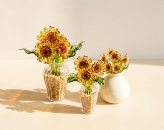 May include: Three glass sunflower arrangements. Two clear, ribbed glass vases are filled with small pebbles and amber-colored glass sunflowers with green stems and leaves. A third, smooth white ceramic vase holds a similar glass sunflower bouquet.