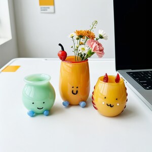 May include: Three small glass vases with cartoon faces. One is light green with blue feet, one is orange with a cherry topper and blue feet, and one is yellow with red horns and a cat-like face. The orange vase holds a bouquet of flowers.