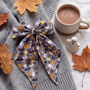 Può includere: Un fiocco per capelli in tessuto grigio con un motivo ripetuto di Halloween composto da zucche arancioni, fantasmi bianchi, pipistrelli neri, caramelle a forma di mais gialle, ossa bianche e la scritta "Boo!" in nero. Il fiocco ha due code lunghe e appuntite.