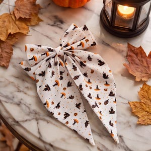 May include: A white fabric hair bow featuring a repeating pattern of orange pumpkins, black witch hats, and dark silhouettes of haunted houses and tombstones, accented with small orange dots across the surface of the bow.
