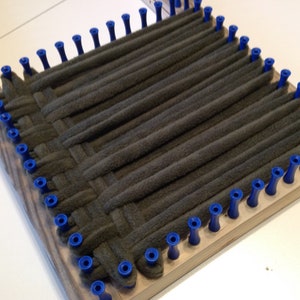Puede incluir: Un telar de madera con clavijas azules y una tela gris tejida a trav&eacute;s de las clavijas.