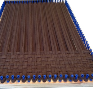 May include: Brown yarn is woven on a wooden loom with blue pegs. The yarn is woven in a simple pattern, creating a textured fabric.