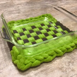 May include: A green and brown crocheted hot pad with a clear glass baking dish on top.
