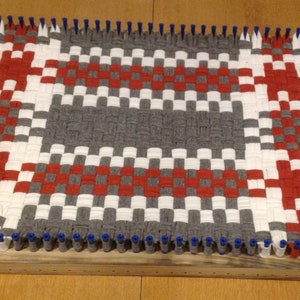 Puede incluir: Una alfombra tejida con un patr&oacute;n de cuadros en rojo, blanco y gris. La alfombra se est&aacute; tejiendo en un telar con clavijas azules.