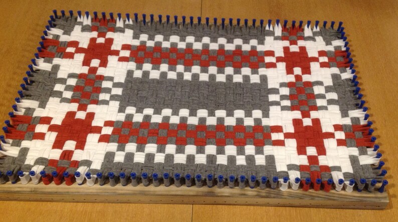 Puede incluir: Una alfombra tejida con un patr&oacute;n de cuadros en rojo, blanco y gris. La alfombra se est&aacute; tejiendo en un telar con clavijas azules.