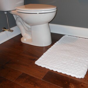 Puede incluir: Una alfombra de ba&ntilde;o blanca con superficie texturizada se encuentra sobre un suelo de madera oscura junto a un inodoro blanco.