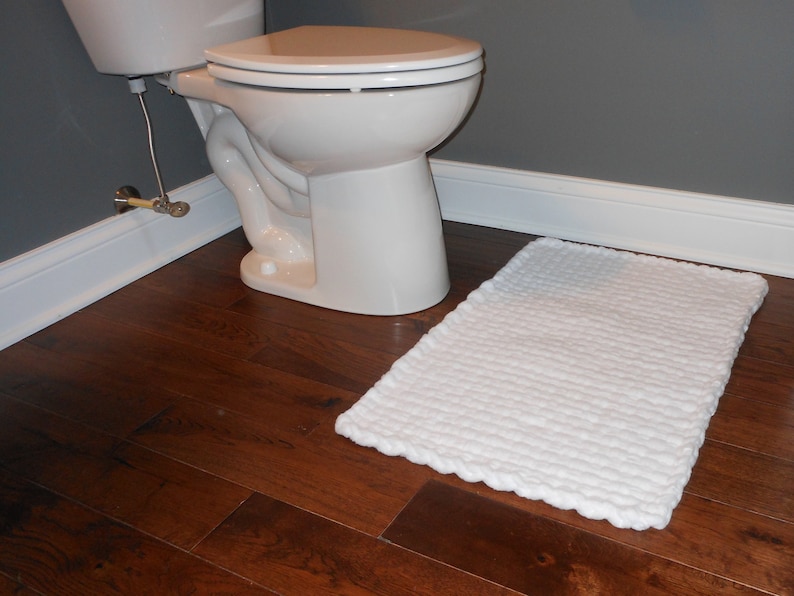 Puede incluir: Una alfombra de ba&ntilde;o blanca con superficie texturizada se encuentra sobre un suelo de madera oscura junto a un inodoro blanco.