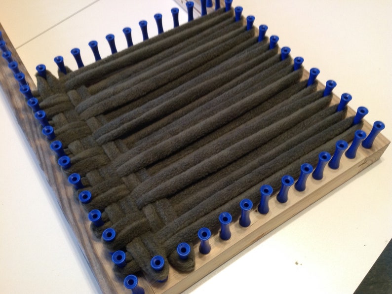 Puede incluir: Un telar de madera con clavijas azules y una tela gris tejida a trav&eacute;s de las clavijas.