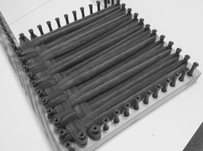 Puede incluir: Primer plano de un telar con hilo negro tejido en un patr&oacute;n cuadrado. El telar est&aacute; hecho de madera y tiene clavijas negras alrededor de los bordes.