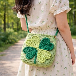 May include: A light green crochet crossbody bag featuring a large four-leaf clover design in shades of green. The bag has a woven texture and a matching green strap. The bag is worn over the shoulder.