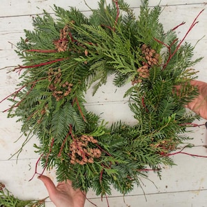 May include: A handmade evergreen wreath with a circular shape. The wreath is composed of green fir branches, accented with small brown pinecones and bright red twigs. The wreath is displayed on a white wooden surface.