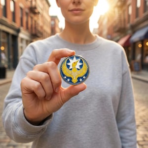 Puede incluir: Una moneda redonda de color dorado con un borde azul. La moneda presenta una estrella blanca, un pájaro amarillo con las alas extendidas y tres estrellas blancas a cada lado. La moneda se sostiene entre los dedos.